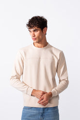 A man wears a plain, cream-colored slim fit round neck sweatshirt with blue jeans.