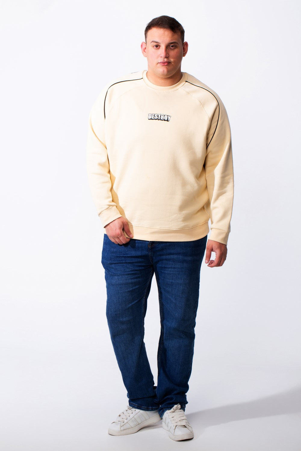 A man wears a cream-colored big size melton sweatshirt with piping detail and blue jeans.
