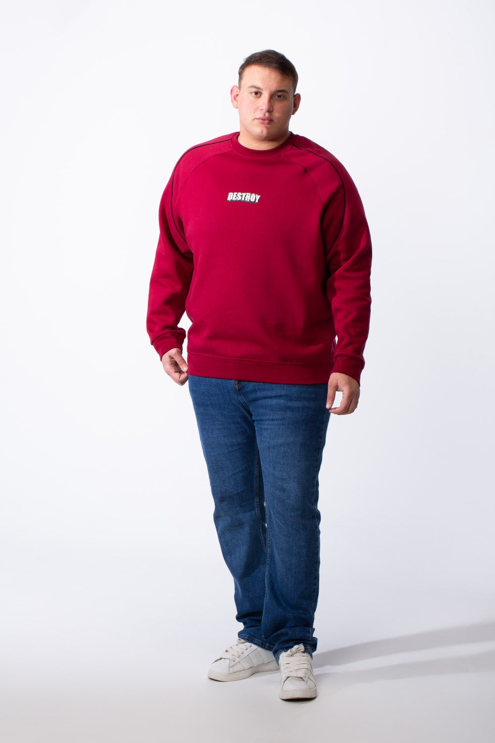 A man wears a big size red crew neck sweatshirt with piping detail and blue jeans.