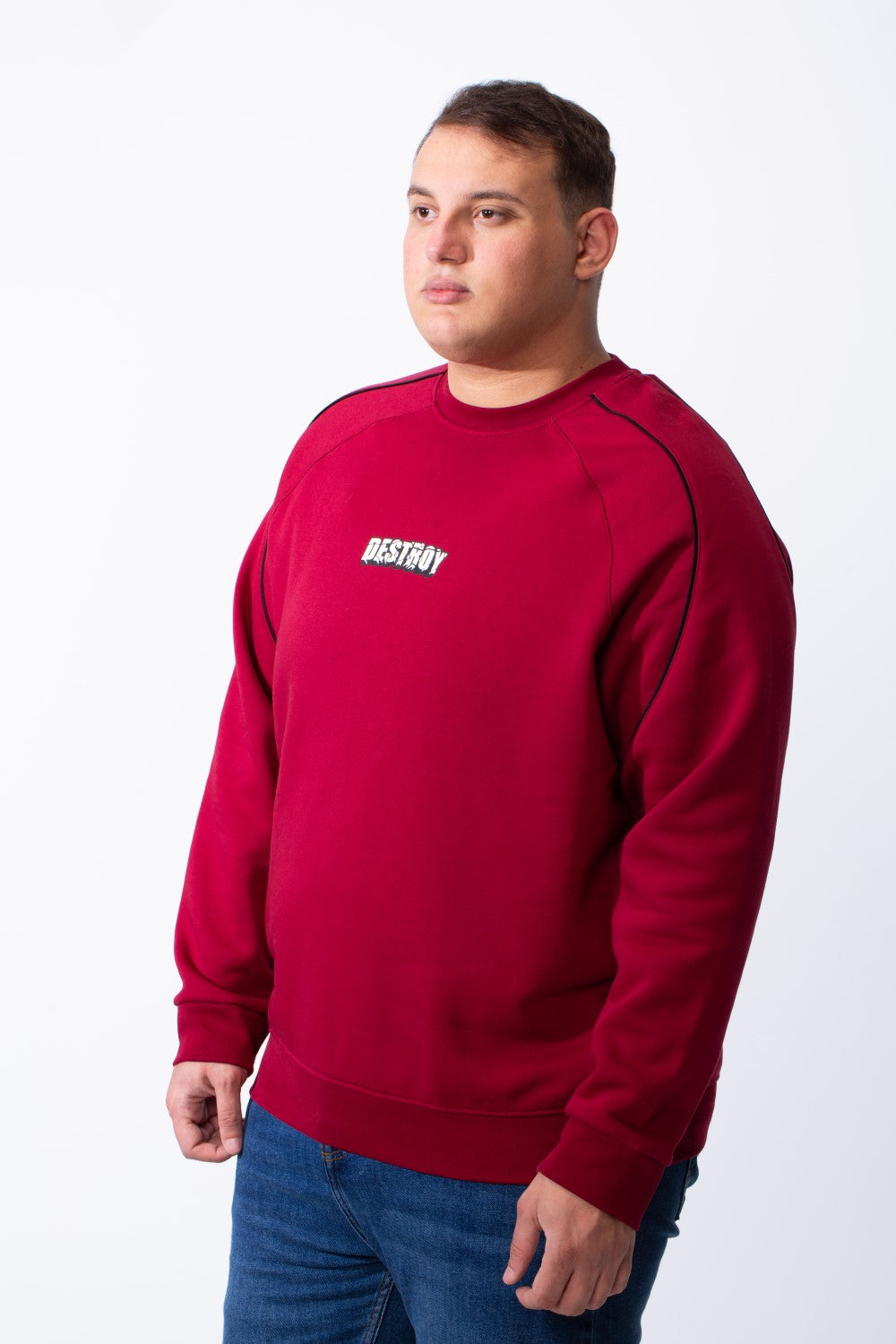 A man wears a big size maroon crew neck sweatshirt with black piping and a small chest graphic.