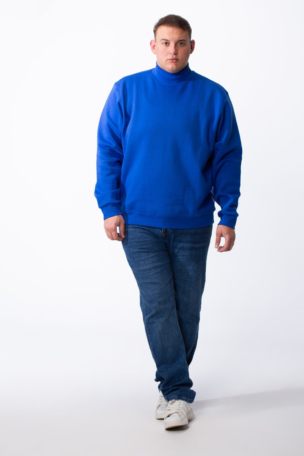 A man wears a blue big size mock neck sweatshirt with ribbed cuffs and dark blue jeans.