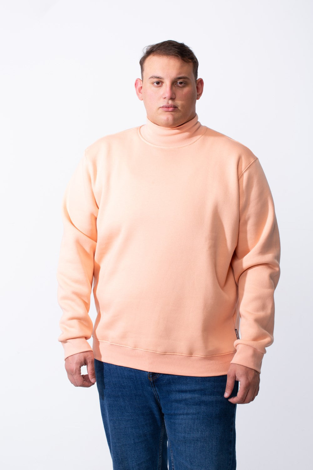 A man wears a peach-colored big size mock neck sweatshirt with ribbed cuffs and blue jeans.