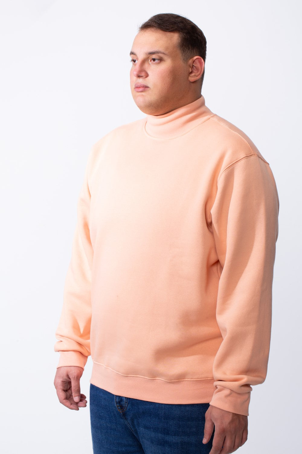 A man wears a peach-colored big size mock neck sweatshirt with ribbed cuffs and blue jeans.