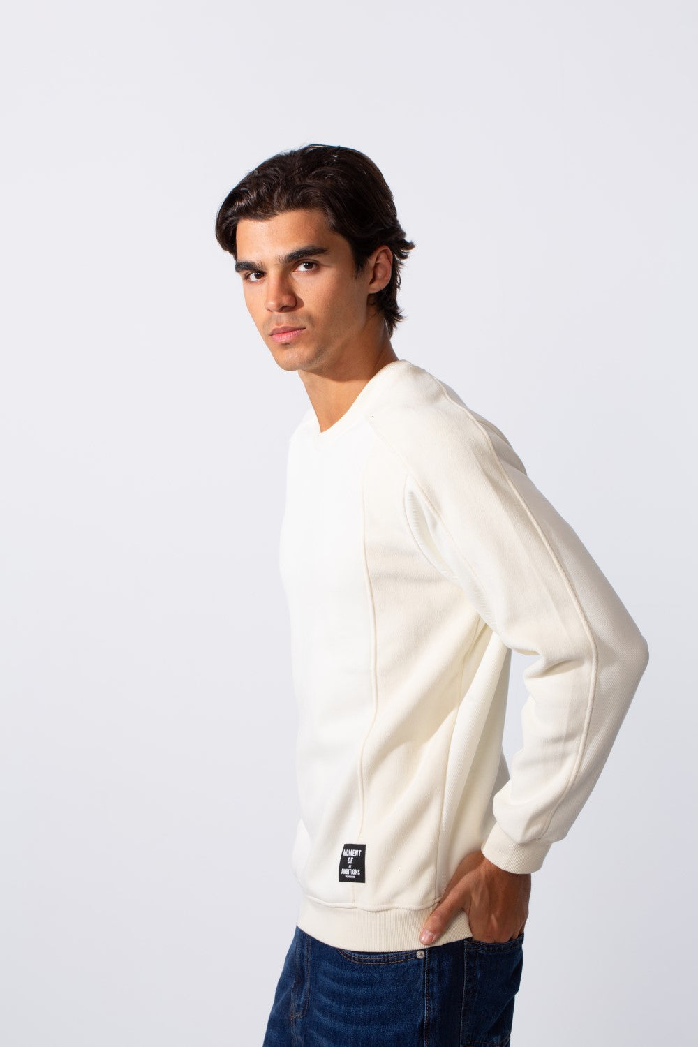 A man wears a slim-fit, off-white round neck sweatshirt with a small black patch and blue jeans.
