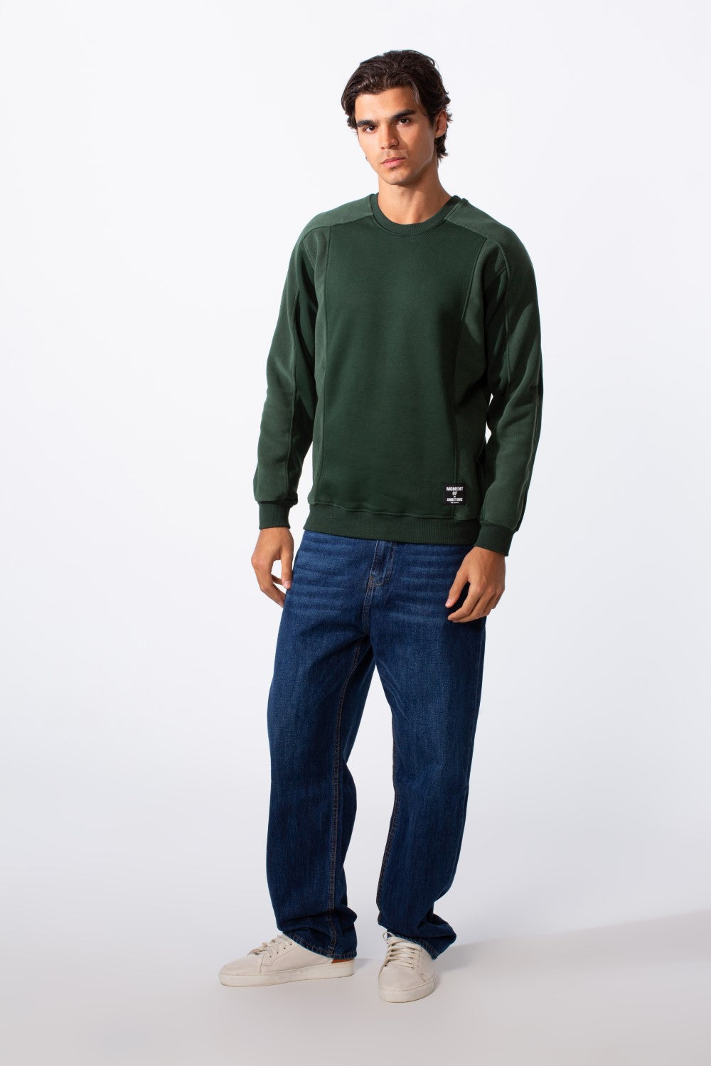 A man wears a dark green slim fit round neck sweatshirt with blue jeans and white sneakers.