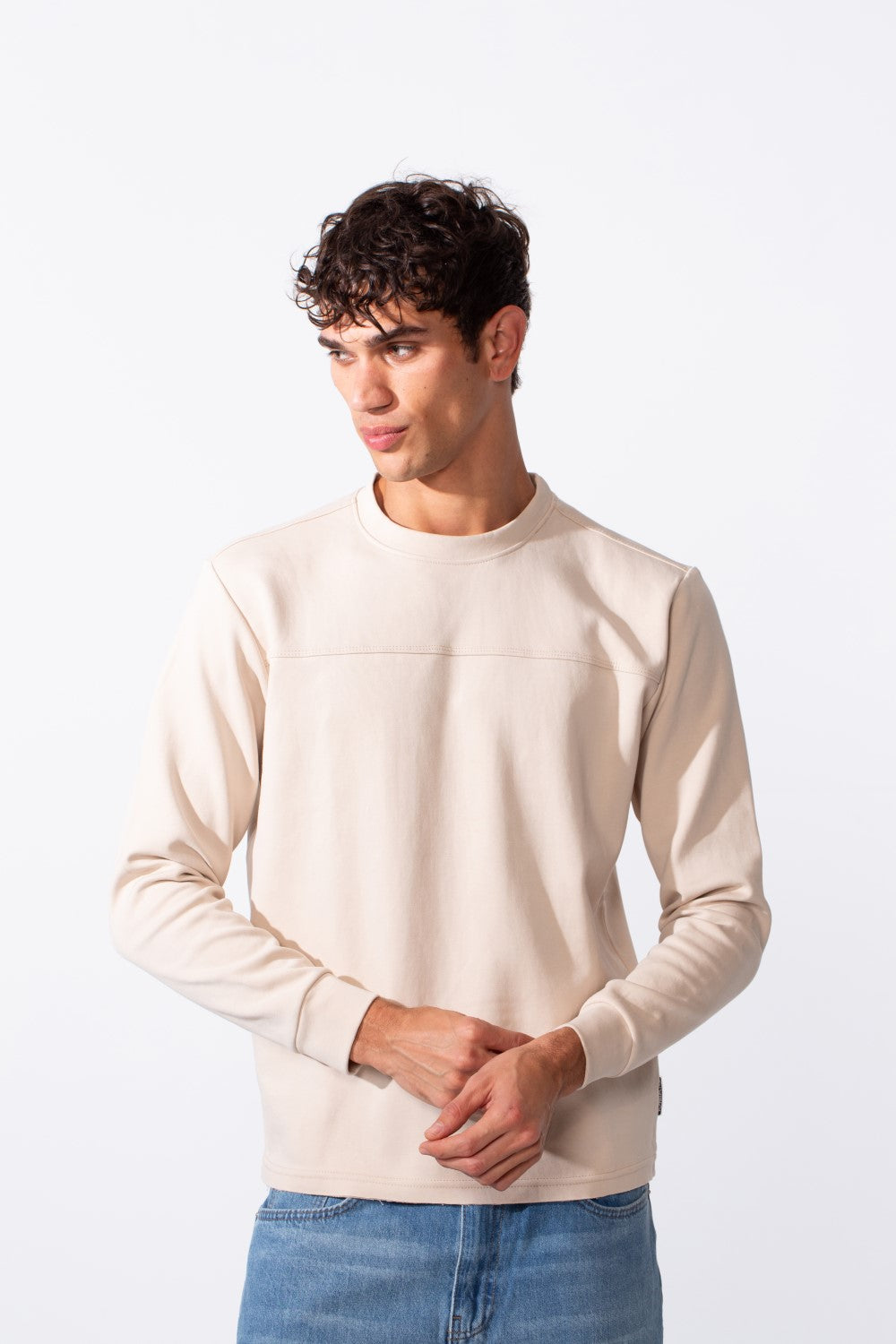 A man wears a plain, cream-colored slim fit round neck sweatshirt with blue jeans.
