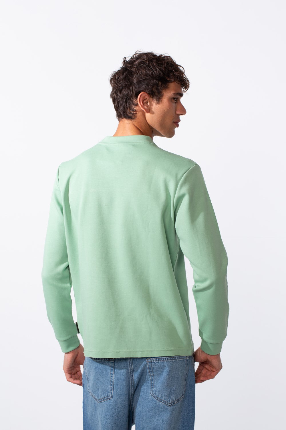 Rear view of a man wearing a light green, slim-fit round neck sweatshirt with blue jeans.