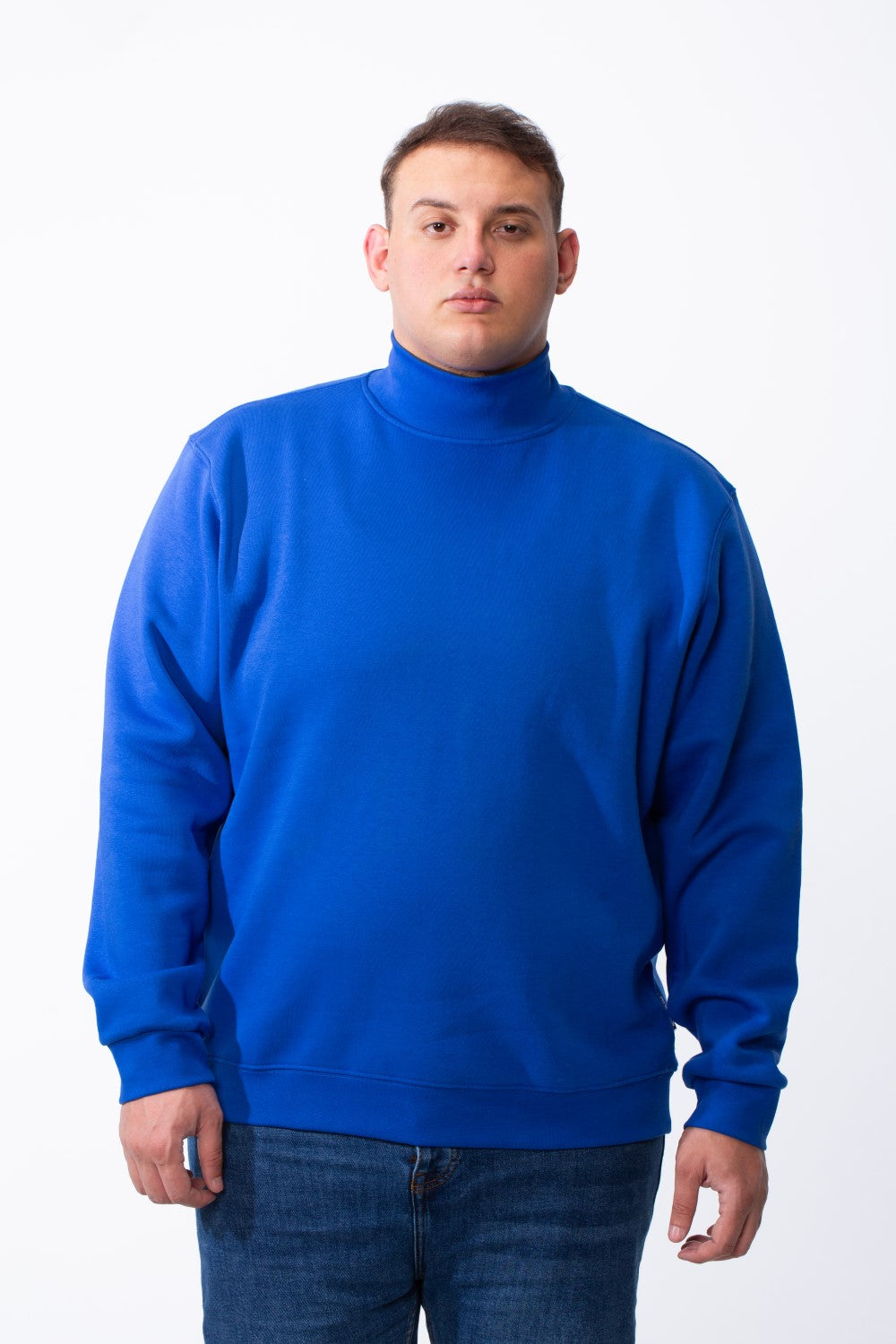 A man wears a royal blue big size mock neck sweatshirt with ribbed cuffs and blue jeans.