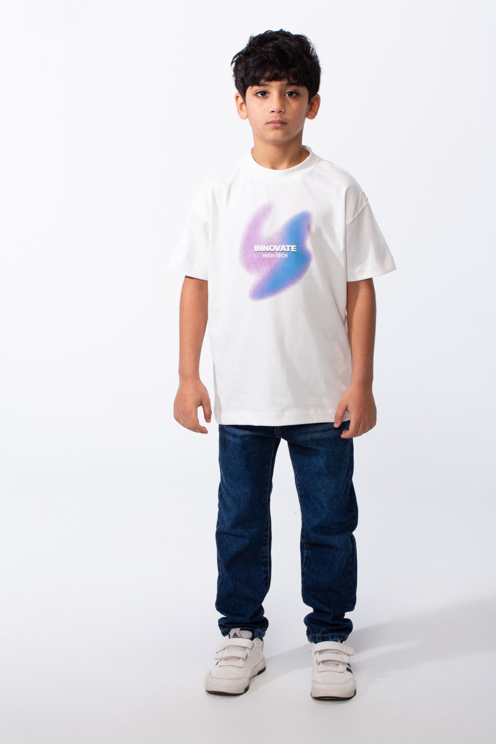 A boy wears a white ribbed crew neck t-shirt with a purple and blue graphic and dark blue jeans.