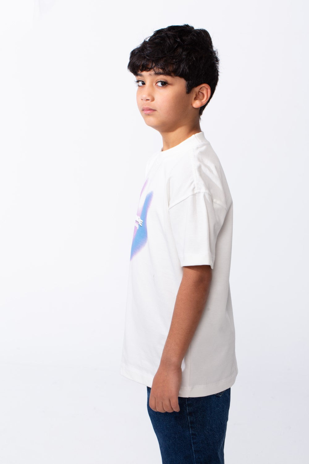 A boy wears a white ribbed crew neck t-shirt with a colorful typography graphic and blue jeans.