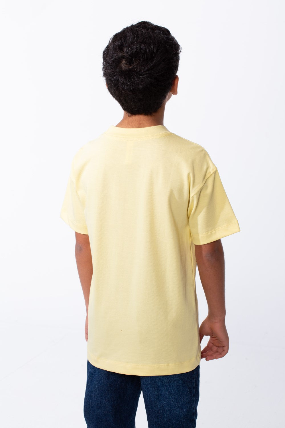 Back view of a boy wearing a pale yellow ribbed crew neck t-shirt and dark blue jeans.