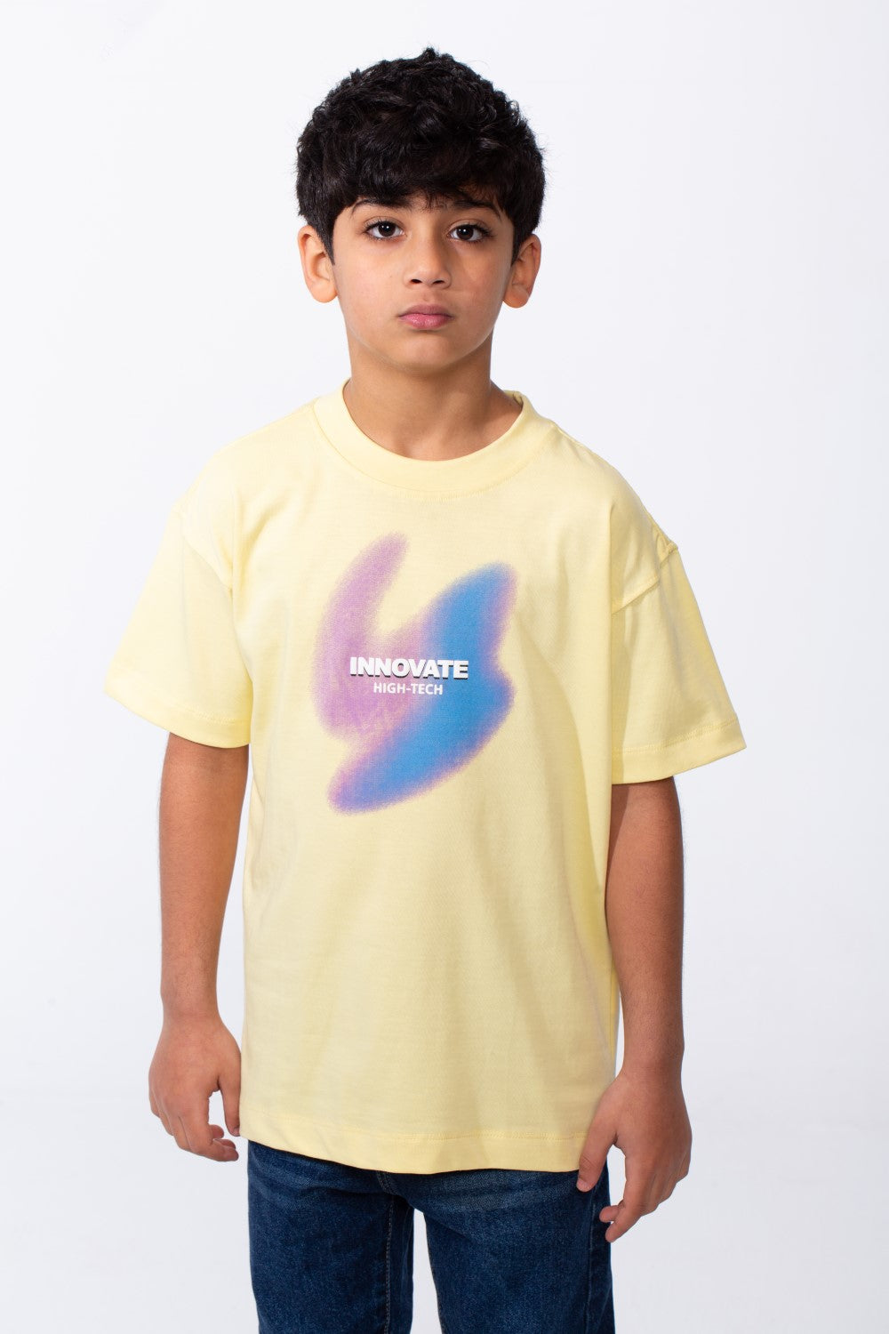 A boy wears a pale yellow crew neck t-shirt with a purple and blue graphic and typography.