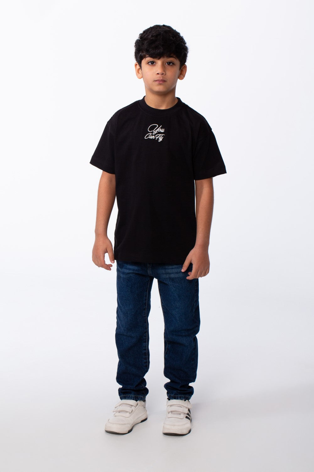 A boy wears a black oversized crew neck t-shirt with white typography embroidery and blue jeans.