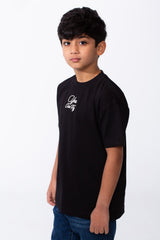 A boy wears a black oversized crew neck t-shirt with white typography embroidery on the chest.